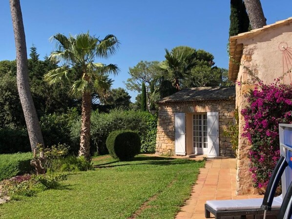 A stone building with a tiled roof and white shutters, surrounded by lush greenery and palm trees.