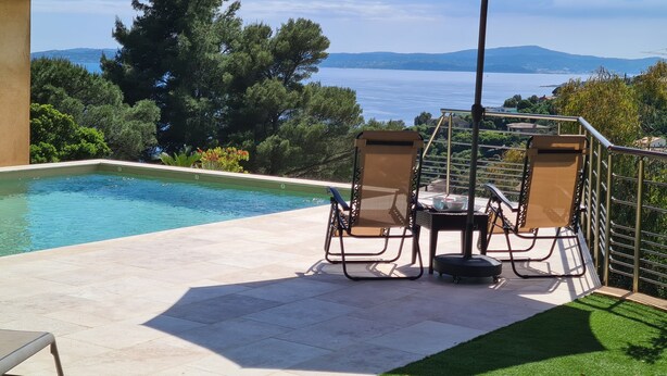 Un espace piscine comprenant deux chaises longues et une table, un parasol rouge, et une vue sur la mer et les collines.