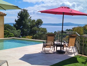 Un espace piscine comprenant deux chaises longues et une table, un parasol rouge, et une vue sur la mer et les collines.