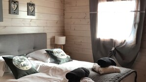 A neatly made bed with a dark quilt, two pillows, and a blanket folded on top. There is a window with curtains, a wall clock, and a ceiling fan.