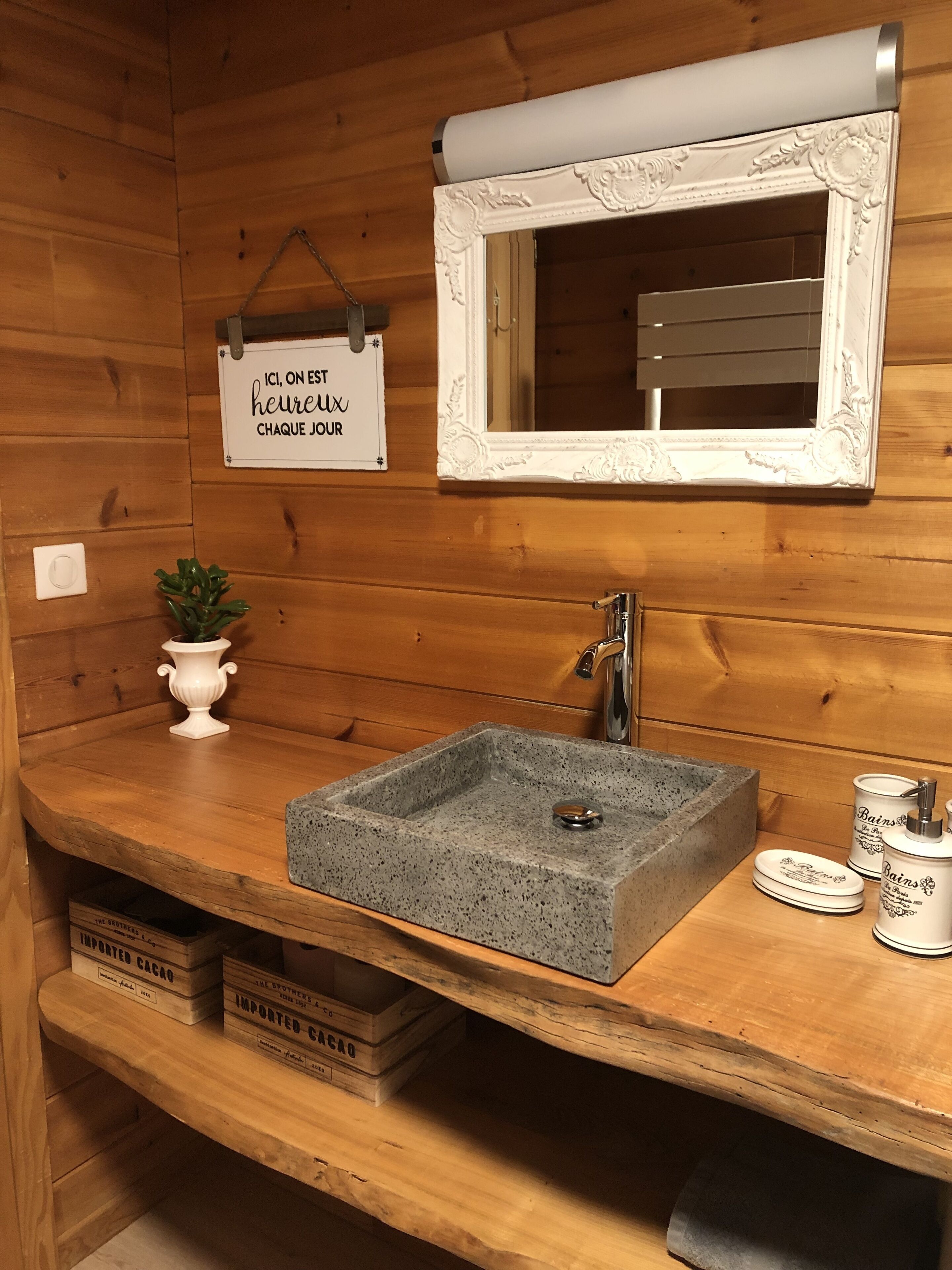 A wooden cabin bathroom with a stone sink, a white vanity, a mirror with an ornate frame, a small potted plant, and a sign above the mirror.