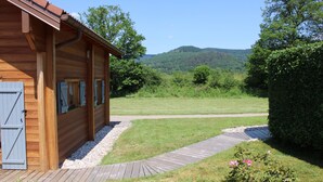 A wooden cabin with a wooden deck, a gravel path, and a view of a green field and hills.