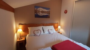 Una cama bien hecha con almohadas con forma de corazón, dos lámparas de noche y un cuadro de un paisaje montañoso en la pared.