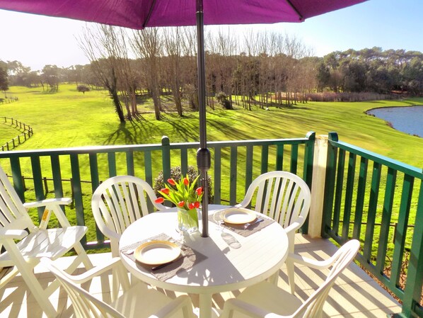 A round table with white chairs and a purple umbrella, set on a balcony overlooking a green landscape with trees and a body of water.