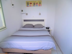 Un lit recouvert d’une couvre-lit à rayures, une table de chevet avec une radio et un mur orné d’une œuvre d’art sur le thème des cactus.