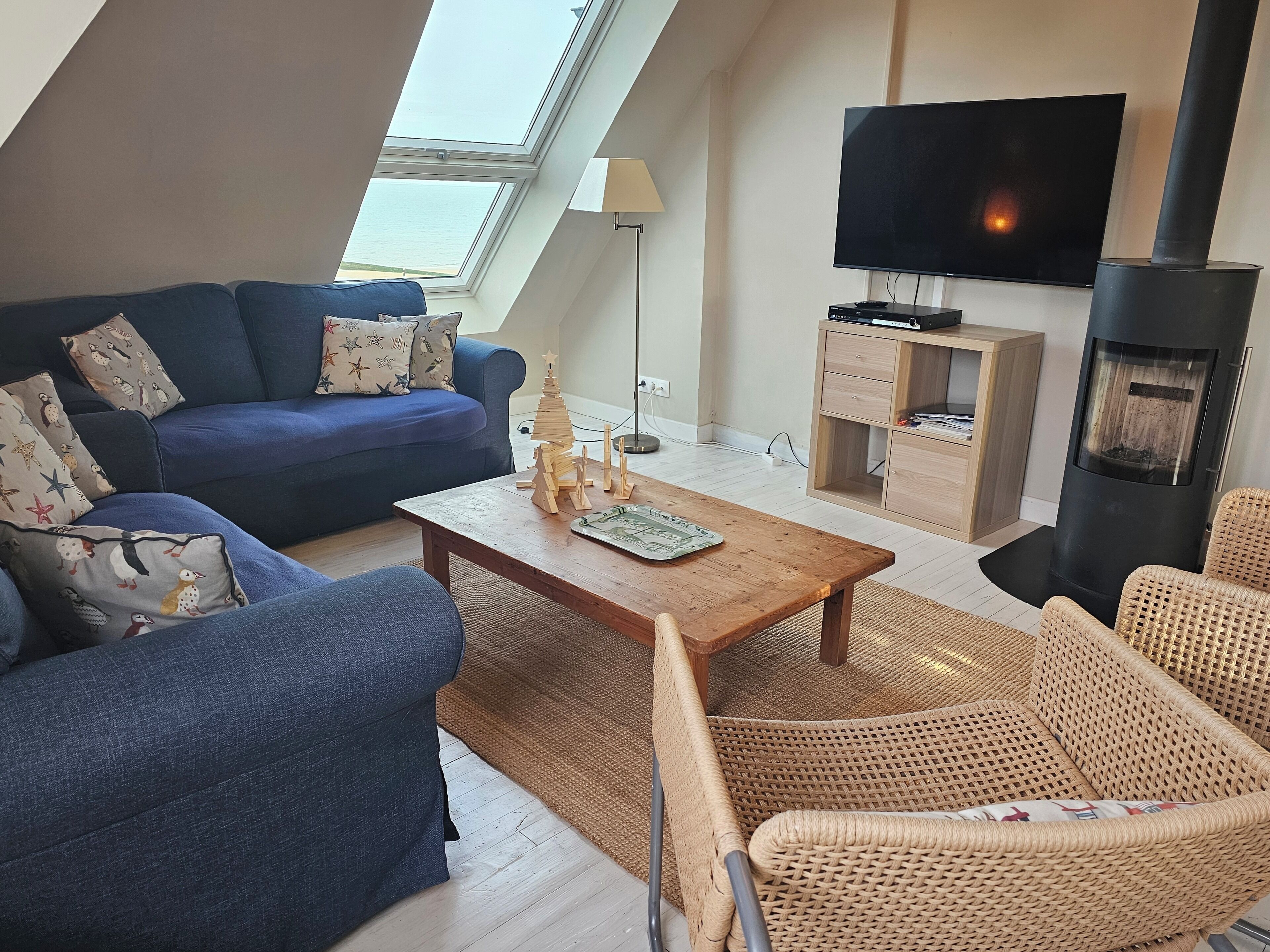 Un salon chaleureux avec un canapé bleu, une table basse en bois, des fauteuils en osier, un téléviseur, une cheminée et une lucarne.