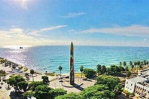 A coastal view with a tall monument, a beach, and a promenade with trees and buildings. - Luxury Apartment at Riviera Colonial (Santo Domingo Este)