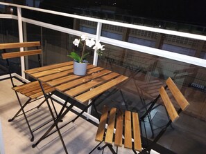 A balcony with a wooden table and chairs, a potted plant, and a glass railing. - Luxury Apartment at Riviera Colonial (Santo Domingo Este)