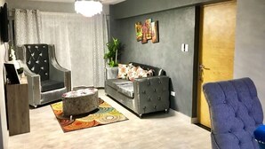 A living room with a grey sofa, a black armchair, a blue armchair, and a round ottoman. There is a ceiling light, a door, and a wall with a framed picture. - Luxury Apartment at Riviera Colonial (Santo Domingo Este)