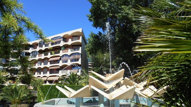 Un hotel de varios pisos con balcones, una fuente de agua y vegetación circundante.