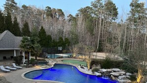 A swimming pool with a basketball court, surrounded by trees and a house.