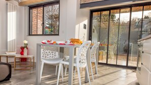 Un coin repas avec une table dressée pour quatre personnes, des chaises blanches et une vue sur l’extérieur, visible à travers une porte vitrée.