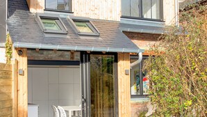 Une maison moderne à deux étages, dotée d’une grande porte d’entrée en verre, d’un coin repas avec des chaises blanches et d’un petit jardin.