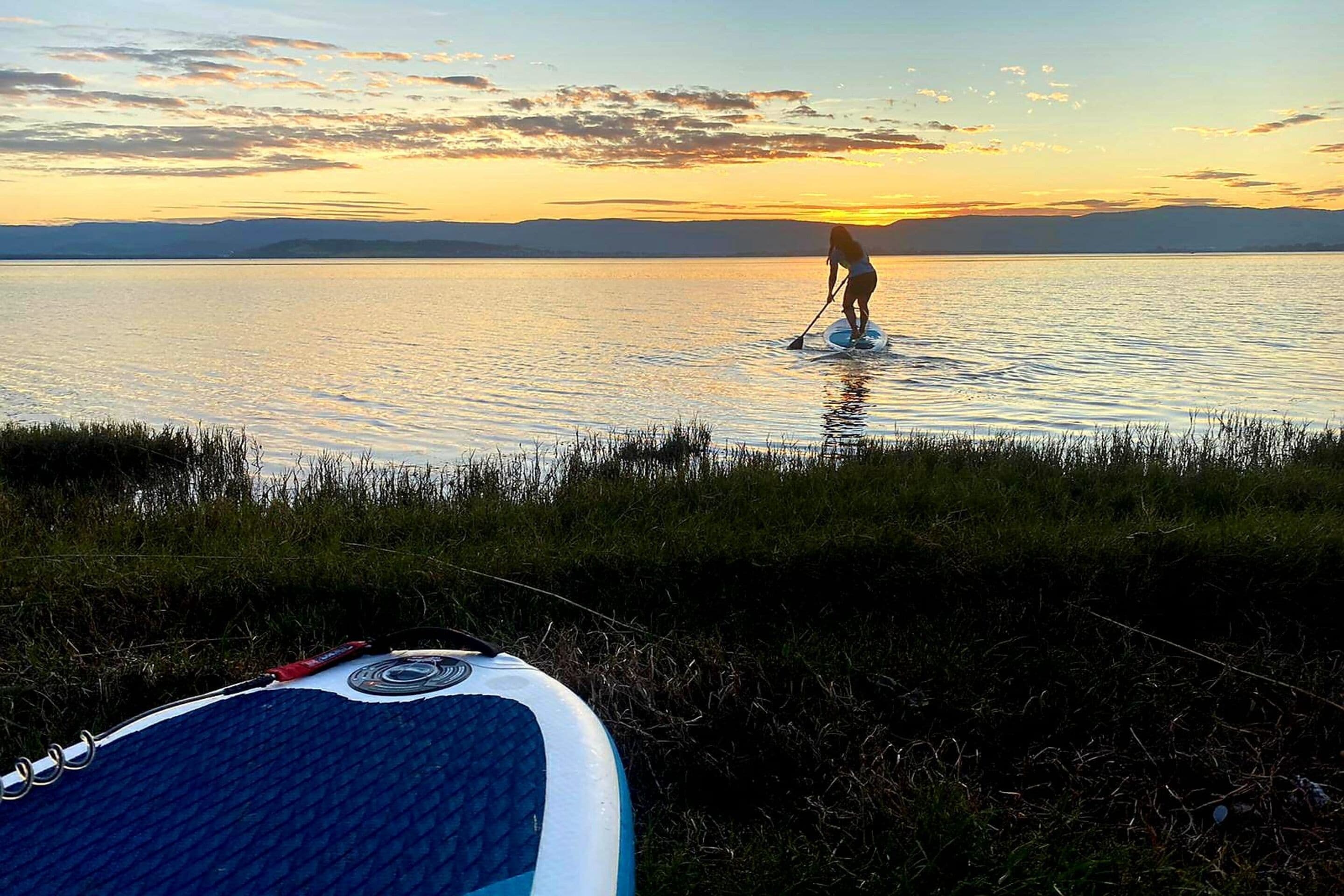 Eine Person steht auf einem Stand-Up-Paddleboard auf ruhigem Wasser bei Sonnenuntergang.