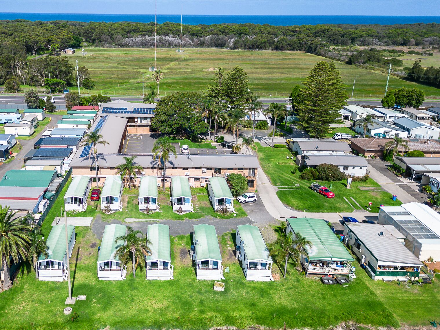 Un camping con vehículos recreativos y casas móviles, rodeado de campos verdes y árboles, con una vista despejada del océano al fondo.