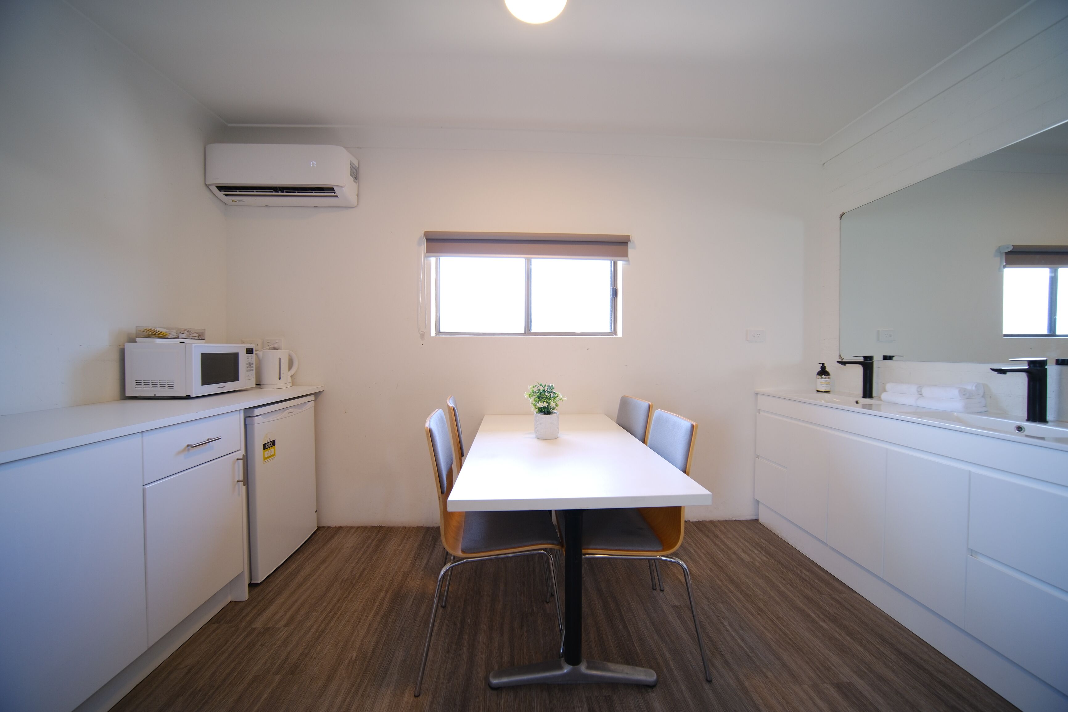 A compact kitchen with a white table, chairs, a microwave, a small refrigerator, and a window with blinds.