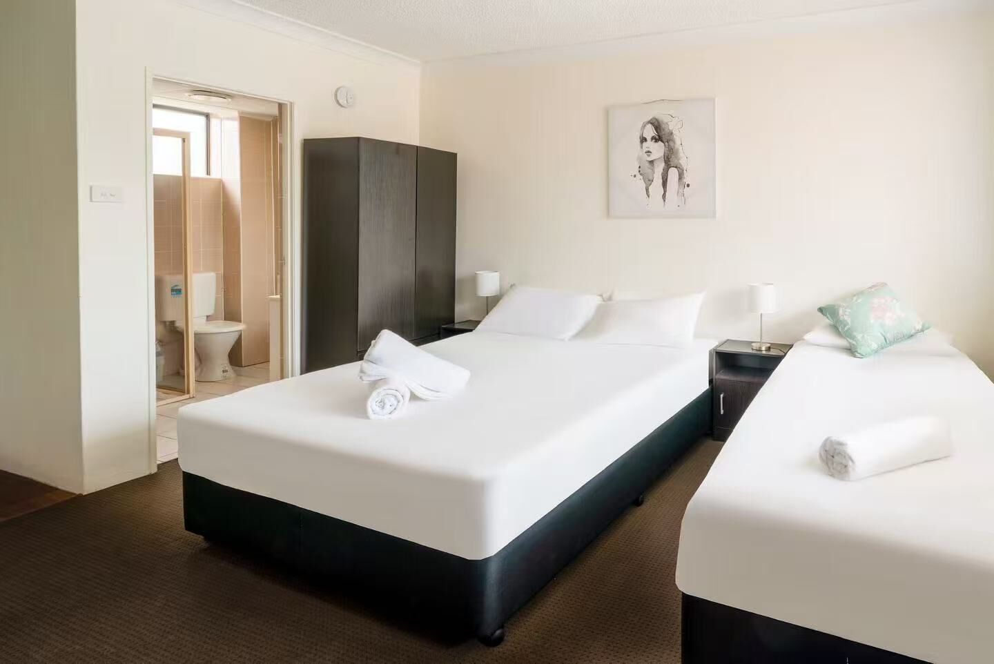 Une chambre d’hôtel avec un grand lit, un plus petit lit, une armoire, une salle de bain avec des toilettes et un tableau au mur.