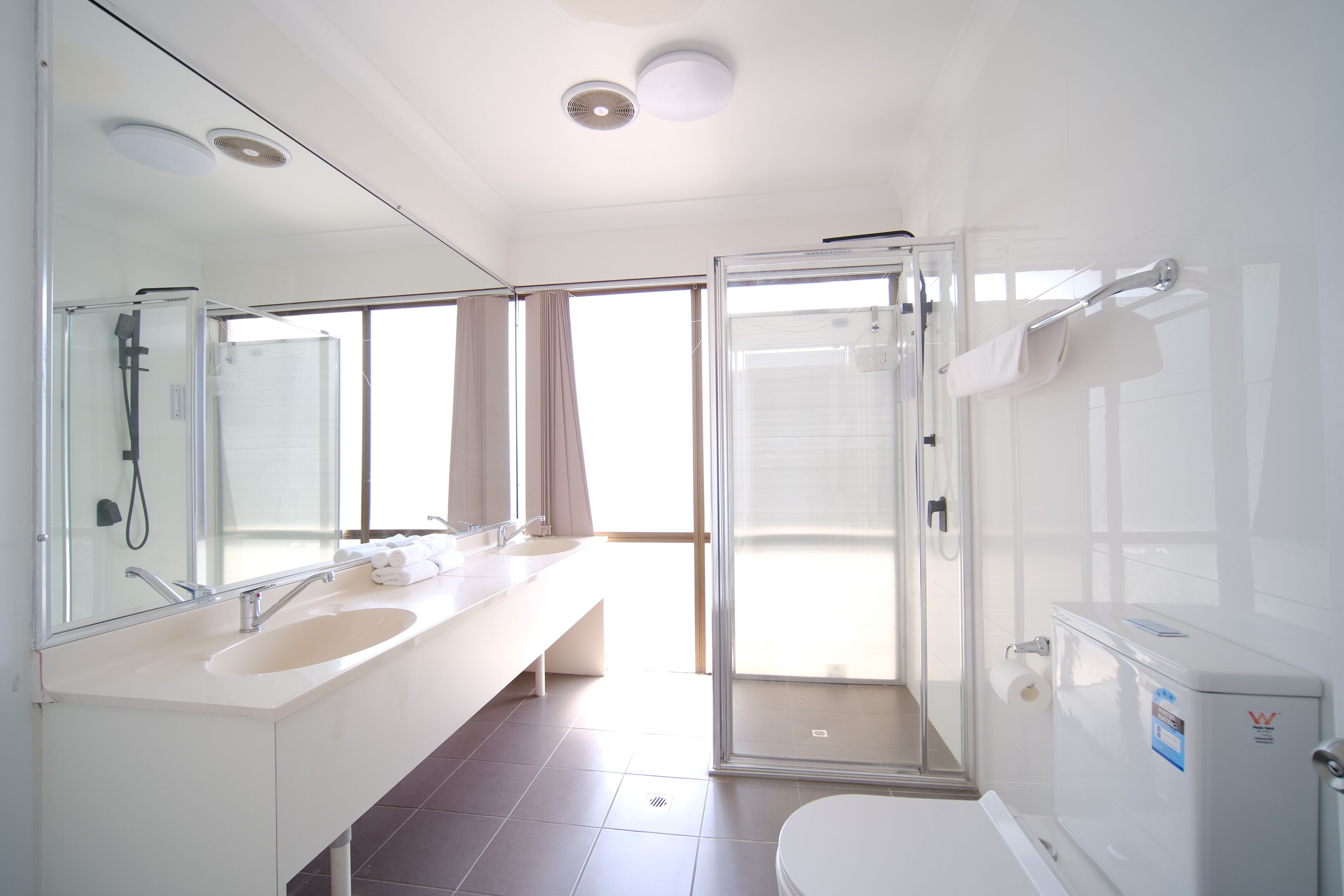 A modern bathroom with a large mirror, two sinks, a shower, and a toilet.