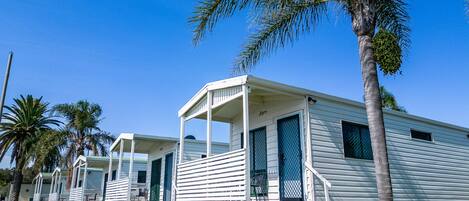 A row of white mobile homes with a porch, surrounded by palm trees and grass.