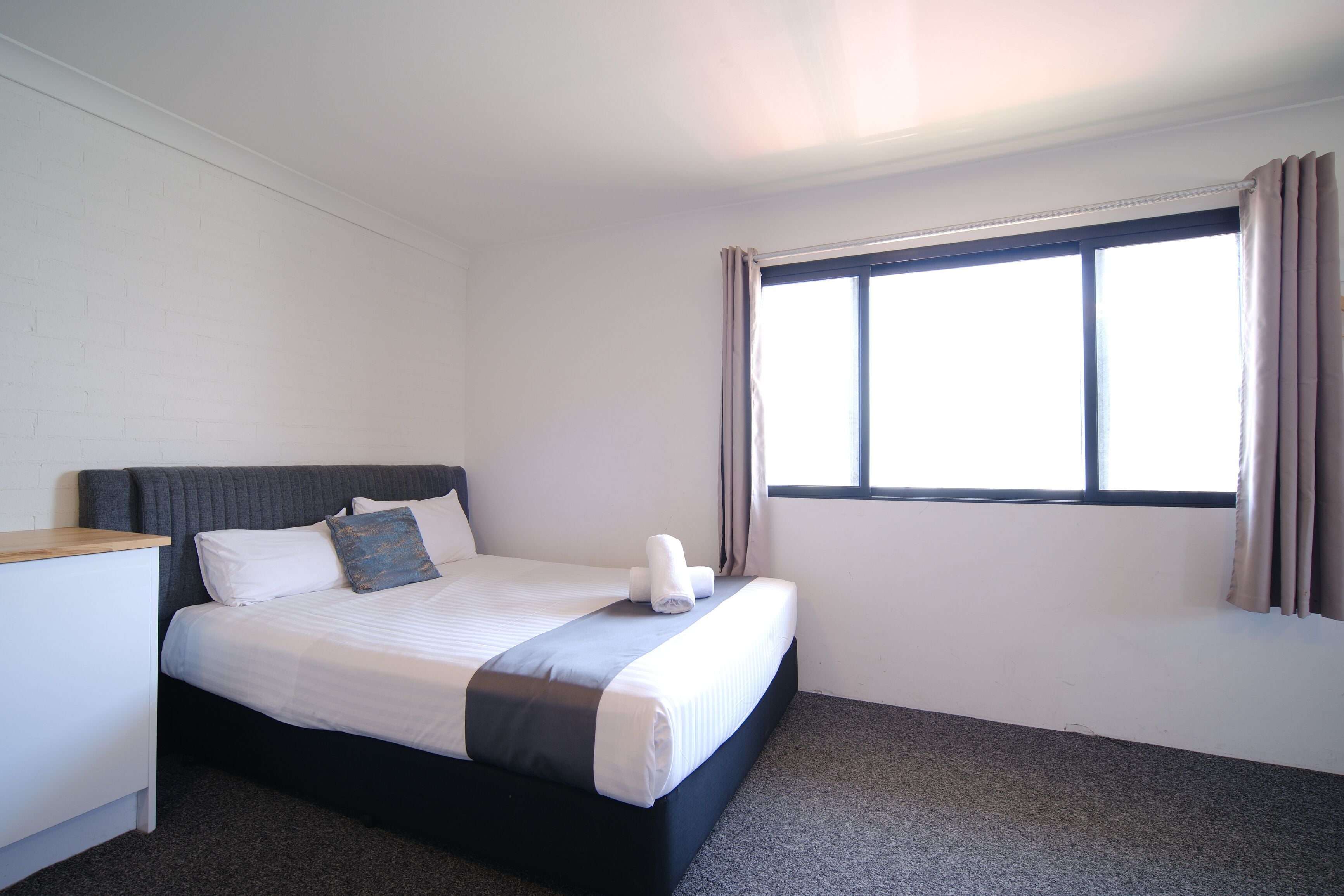 A neatly made bed with a gray headboard, white bedding, and a blue pillow. There is a small bedside table with a white lamp. The room has a large window with beige curtains, and the walls are white.
