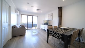 A modern living room with a sofa, a dining table with chairs, a TV, and a large window. - Lakeside Inn Illawarra  (Windang)
