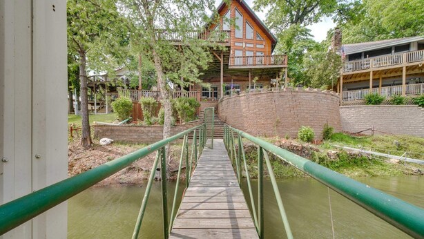 Un puente de madera cruza un canal de agua que lleva a una cabaña con balcón y pared de piedra.