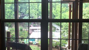 Vista a través de una ventana que muestra una terraza de madera y un pequeño puente sobre un estanque, rodeado de árboles.