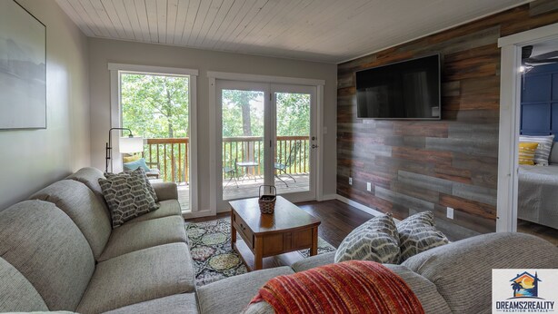 A spacious living room with a large sectional sofa, a wooden coffee table, and a TV mounted on a wooden wall. There is a sliding glass door leading to an outdoor deck with a table and chairs. - Hidden Hideaway | 4BR Connected Condo Near Silver Dollar City (HH9-3+4) (Reeds Spring)