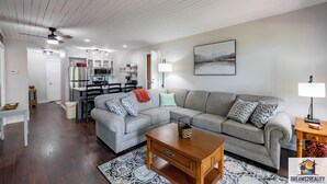 A modern living room with a grey sectional sofa, a wooden coffee table, and a patterned rug. A kitchen is visible in the background with a dining area. - Hidden Hideaway | 4BR Connected Condo Near Silver Dollar City (HH9-3+4) (Reeds Spring)