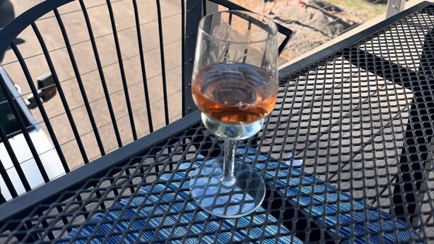 A glass of amber liquid on a metal table with a blue patterned tablecloth, overlooking a forested area with a lake and a paved path. - Lake Almanor West - Lakefront Cabin (Chester)