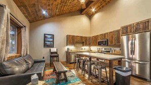 A modern kitchen with wooden cabinets, stainless steel appliances, a bar area, and a cozy seating area with a leather couch and a colorful rug. - Your Basecamp to Glacier• Cozy 2BR w/ King + Queen (Coram)