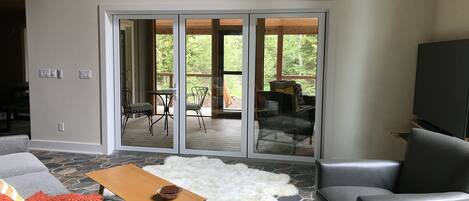 Ein Wohnzimmer mit Steinboden, einem Holztisch, einem grauen Sofa, einem weißen, flauschigen Teppich und einer Glasschiebetür, die auf eine Terrasse mit Stühlen und einem Fernseher führt.