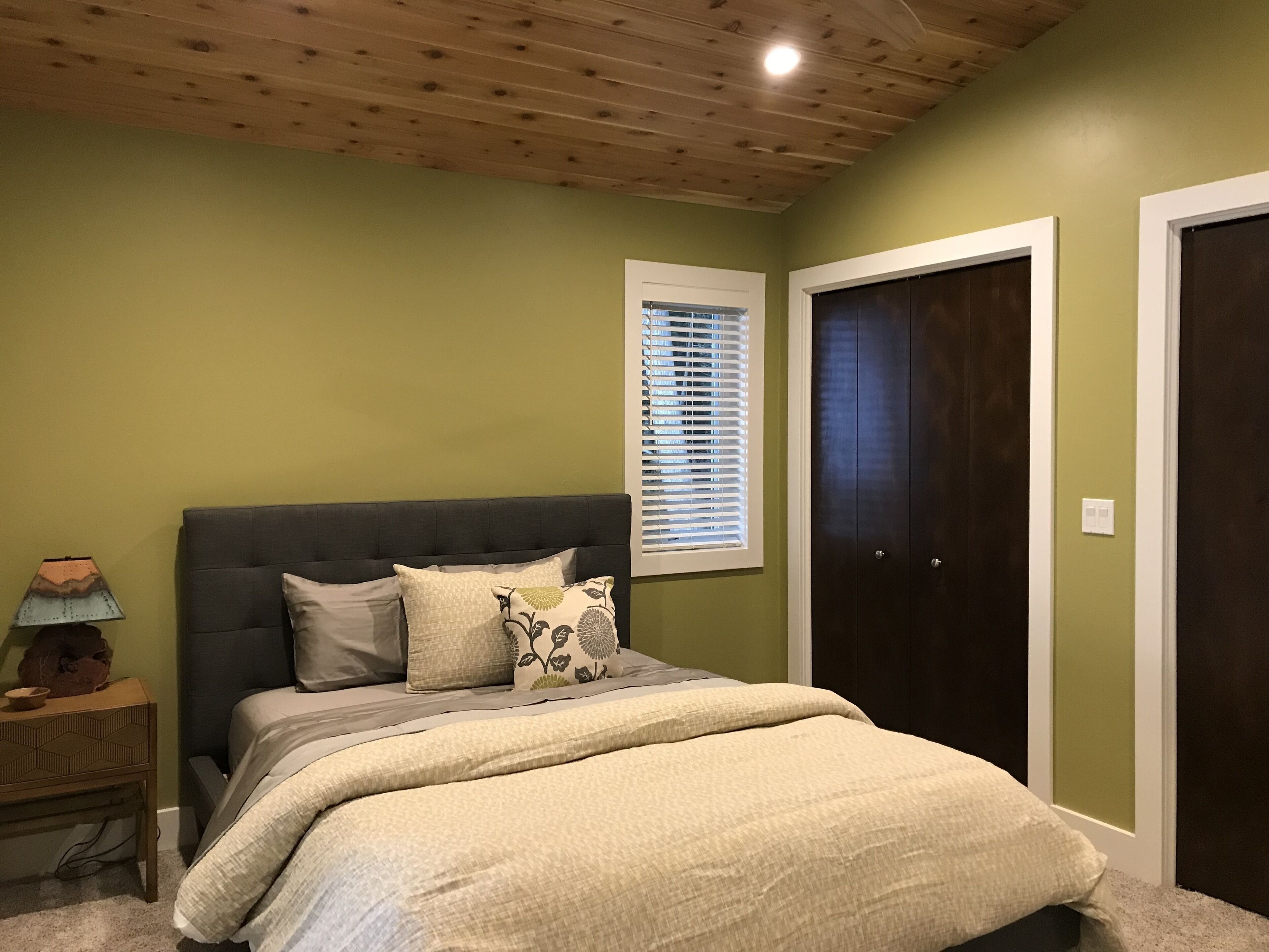 Une chambre à coucher comprenant un lit, une table de chevet, une lampe, une fenêtre avec des stores et une porte.
