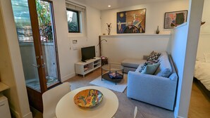 A modern living room with a grey sofa, a white TV stand, a glass coffee table, and a dining table with a colorful bowl. - Ocean & Lagoon View Studio, Walk to Beach, Large Private View Deck  (Del Mar)