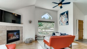 A modern living room with a fireplace, a ceiling fan, and large windows offering a view of trees. - One of the nicest house rentals available in Lyons, Colorado (Lyons)