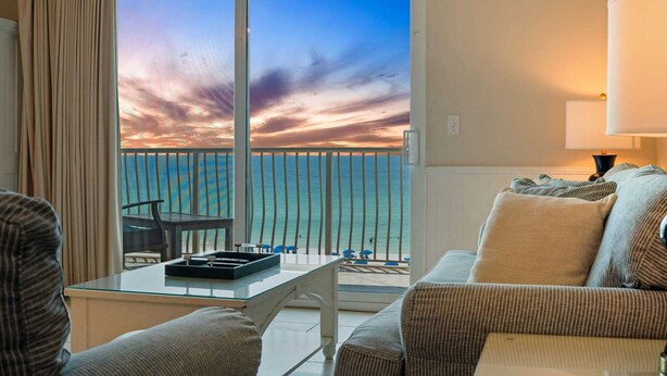 A balcony with a view of the ocean and a sunset. There is a glass table with a tray and a chair. A sofa and a lamp are also visible. - Beach Retreat 407  Gulf Front, Dog friendly, Beach Service (Miramar Beach)