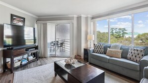 Una sala moderna con televisor de pantalla plana, mueble de entretenimiento de madera oscura, sofá gris con cojines estampados y vista a un balcón y paisaje natural.