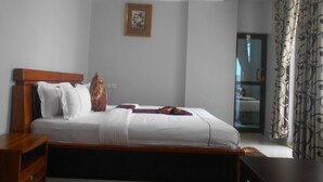 Ein Schlafzimmer mit einem hölzernen Bett, weißer Bettwäsche, einem kleinen Nachttisch, einem gerahmten Bild an der Wand und einem fensterverschönenden, gemusterten Vorhang.