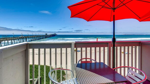 Un balcón con una sombrilla roja, una mesa de metal y sillas con vista a la playa y el muelle.