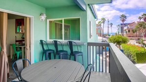 Uma varanda com mesa e cadeiras, uma janela com persianas e vista para o oceano e outros edifícios.