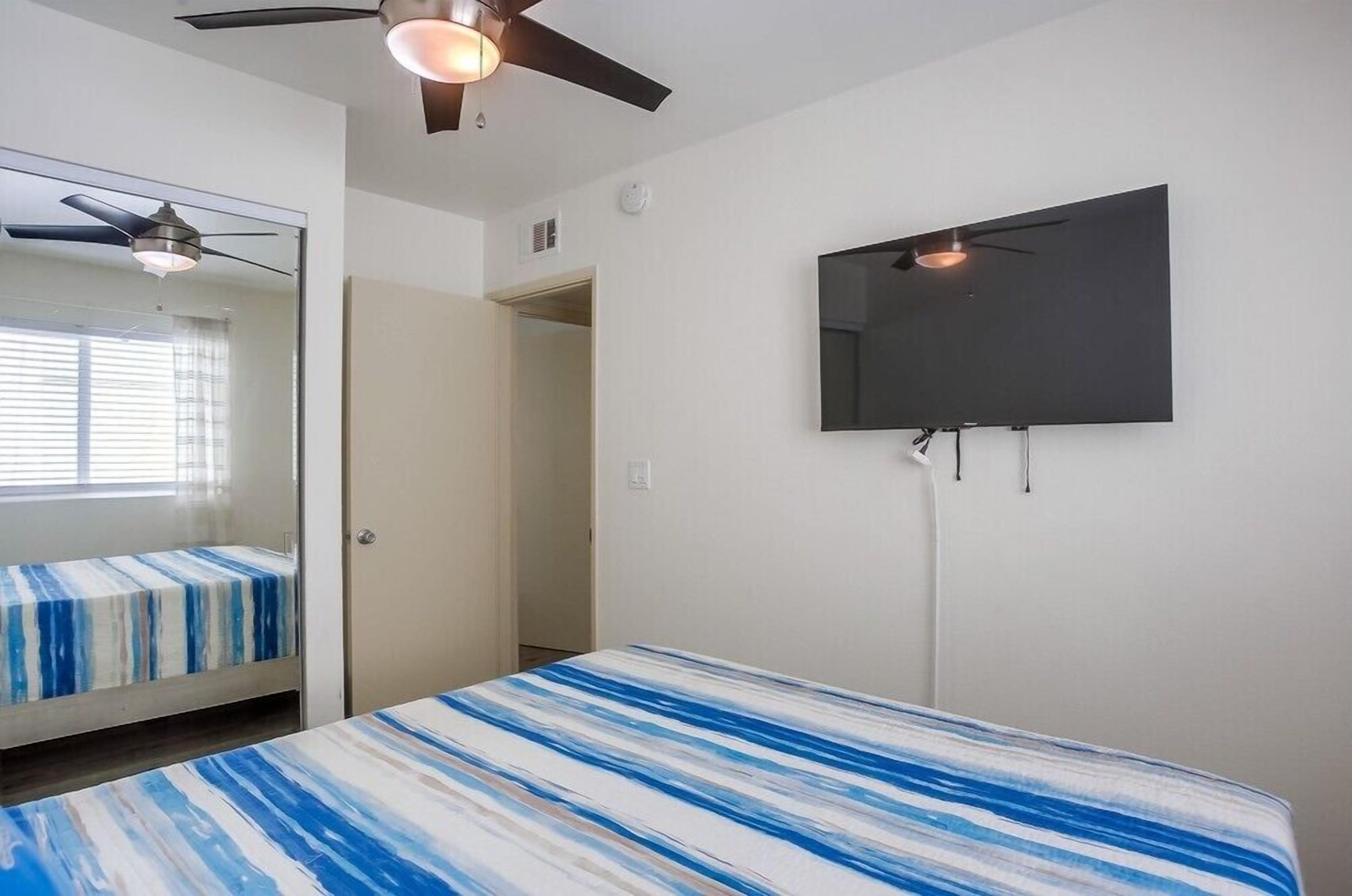 Un dormitorio con cama, ventilador de techo, televisor montado en la pared y una puerta que da a otra habitación.