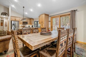 A spacious dining area with a large wooden table and chairs, a kitchen with modern appliances, and a large window with a view of trees.