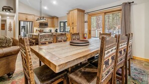 A spacious dining area with a large wooden table and chairs, a kitchen with modern appliances, and a view of the outdoors through a large window. - Walk to Summit, Spa, BBQ, Fireplace (Big Bear)
