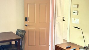 A room with a wooden door, a table, a chair, a lamp, and a remote control.