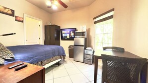 A room with a bed, a desk with a chair, a television, a refrigerator, and a window with blinds.