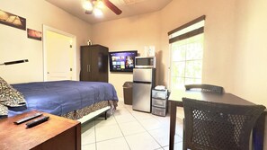 Een kamer met een bed, een bureau met stoel, een televisie, een koelkast en een raam met jaloezieën.