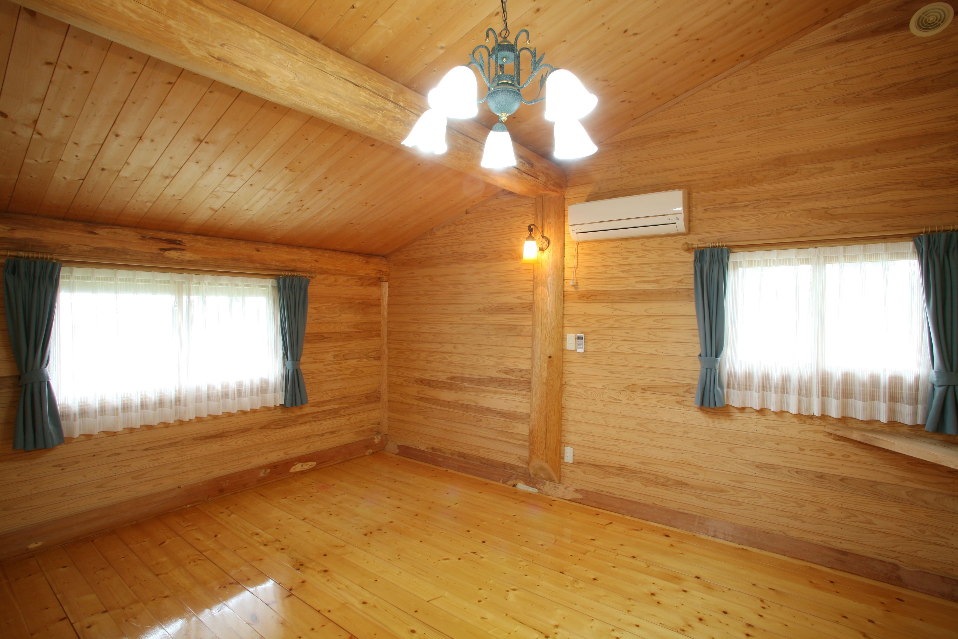 A spacious room with wooden walls and floors, a ceiling fan, and two windows with curtains.