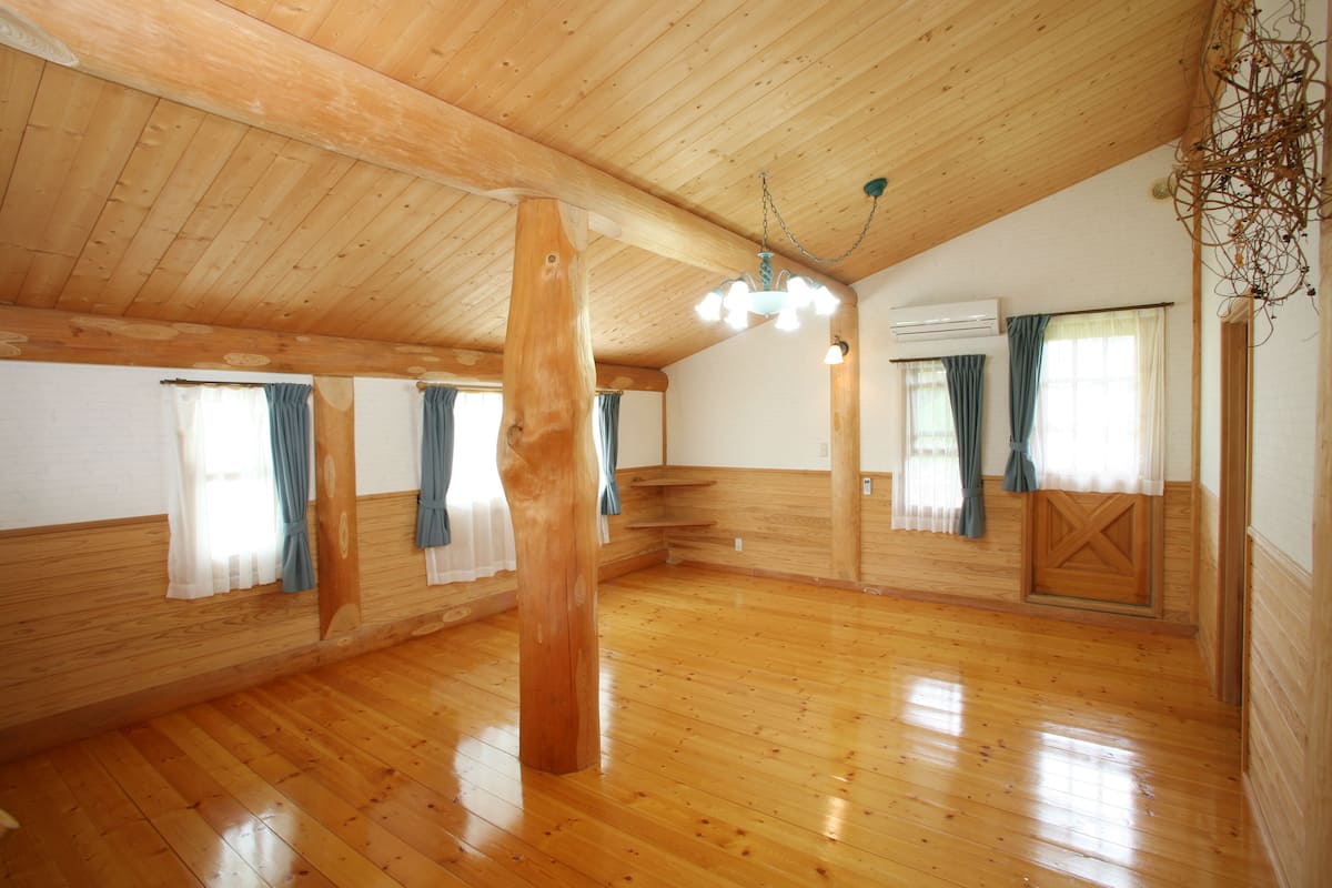 A spacious room with wooden floors and walls, a wooden ceiling, and a chandelier. There are windows with curtains and an air conditioning unit.