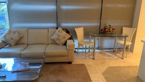 A living room with a sofa, a glass coffee table, a dining table with chairs, and a large window with blinds. - Old Town Scottsdale - Optima Camelview - Modern/Furnished, Walk To All Amenities (Scottsdale)