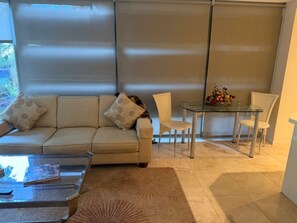 A living room with a sofa, a glass coffee table, a dining table with chairs, and a large window with blinds.